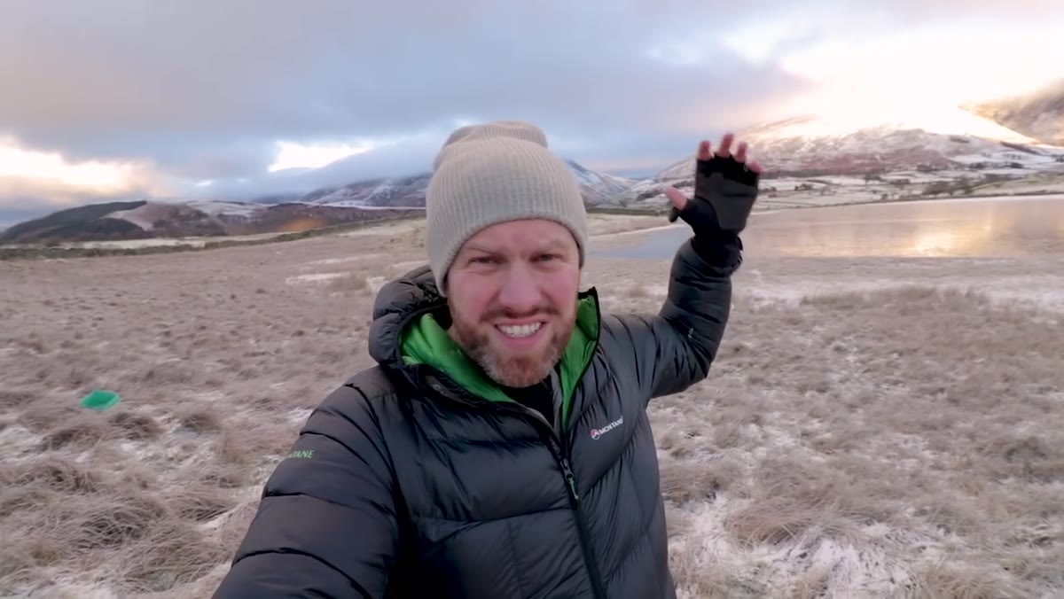 Photographer gesturing across frozen tarn toward distant mountains